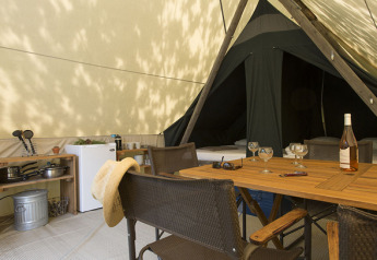 Innenansicht eines Safari-Zeltes Canadienne mit Holztisch, Stühlen, Kühlschrank und Kochutensilien.