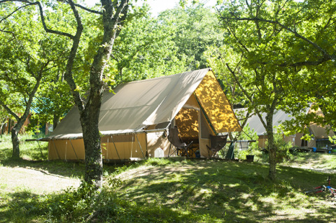 Safari tent named Canadienne tent on a wooded campsite, surrounded by green trees and outdoor chairs.