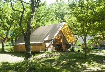 Tenda safari chiamata Canadienne tent in un campeggio boscoso, circondata da alberi verdi e sedie da esterno.