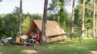 A family sits outside a Canadienne safari tent surrounded by tall trees on a grassy campsite.