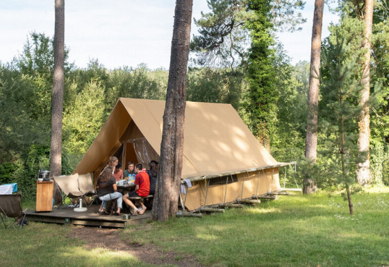 A family sits outside a Canadienne safari tent surrounded by tall trees on a grassy campsite.