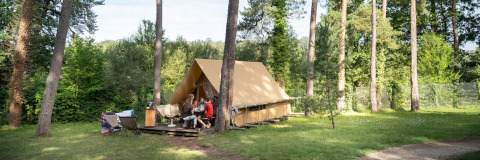 A family sits outside a Canadienne safari tent surrounded by tall trees on a grassy campsite.