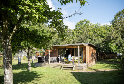 Chalet Portland i dagslys med gæster på terrassen, omgivet af frodige grønne træer og natur.