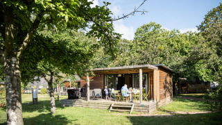 Chalet Portland in het zonlicht met gasten op het terras, omgeven door groene bomen en natuur.