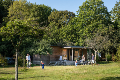 Familie nyder udendørs tid foran Chalet Portland ved Huttopia Parque de Doñana, omgivet af grønne træer.
