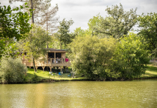 Træhytte kaldet Chalet Portland ved søbredden i Huttopia Parque de Doñana, Spanien, omgivet af frodige træer.