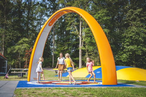 Niños jugando bajo un gran arco naranja en Camping de Koeksebelt en los Países Bajos, rodeados de árboles.