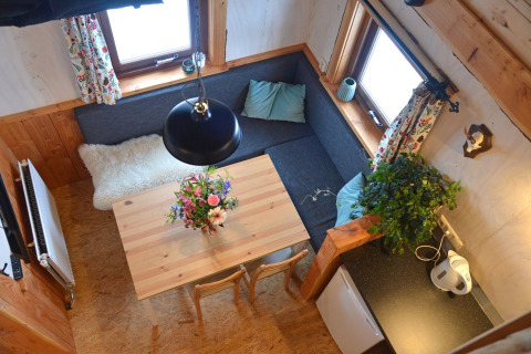 Fugleperspektiv af hyggelig opholdsstue med hjørnesofa, spisebord, blomster og vinduer i et træhus.