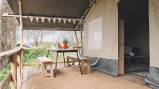 Safari-Zelt-Terrasse mit Holztisch und Bänken, Outdoor-Essen unter Vordach, grüne Umgebung.