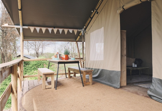 Safari-Zelt-Terrasse mit Holztisch und Bänken, Outdoor-Essen unter Vordach, grüne Umgebung.