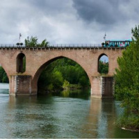Puente de piedra sobre un río con árboles verdes y cielo nublado en un parque de vacaciones de glamping.