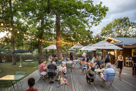 Terraza exterior en Huttopia Lac de l'Uby - Gers, Francia, con personas relajándose bajo luces colgantes.