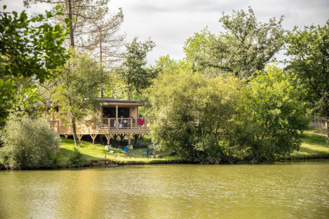 Cabin with deck beside a peaceful lake at Huttopia Lac de l'Uby, Gers, surrounded by lush green trees.