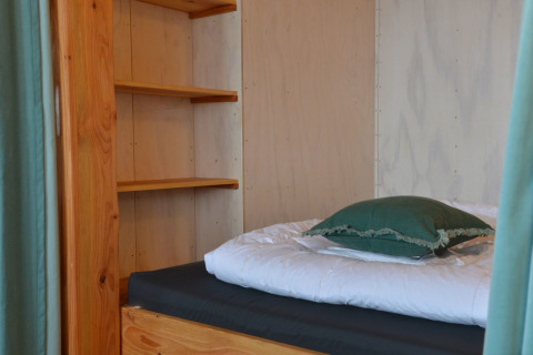 Vista interior de una casa del árbol con cama individual, estanterías y almohada en Camping de Koeksebelt, Países Bajos.