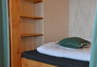 Vista interior de una casa del árbol con cama individual, estanterías y almohada en Camping de Koeksebelt, Países Bajos.