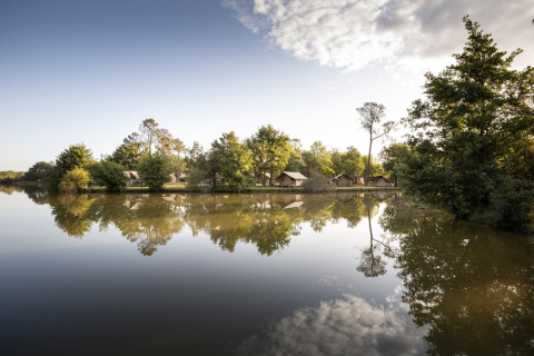 Reflection of trees and small cabins on a lake at Huttopia Lac de l'Uby - Gers, a holiday park in Occitanie.