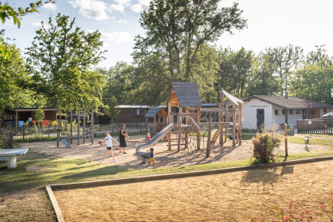 Parque infantil con niños y adultos en Huttopia Lac de l'Uby, Occitania, Francia, rodeado de cabañas de madera.