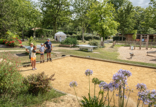 Family playing pétanque at a holiday park in Occitanie, France, with greenery and playground nearby.