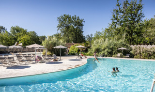 Piscina exterior en Huttopia Lac de l'Uby - Gers, rodeada de tumbonas, sombrillas y abundante vegetación.