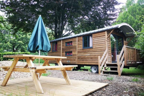 Cabaña Gypsy caravan con terraza de madera, mesa de picnic y sombrilla rodeada de vegetación.
