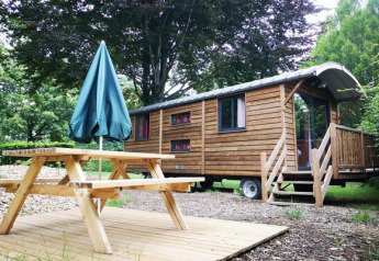 Cabina Gypsy caravan con terrazza in legno, tavolo da picnic e ombrellone immersa nel verde.