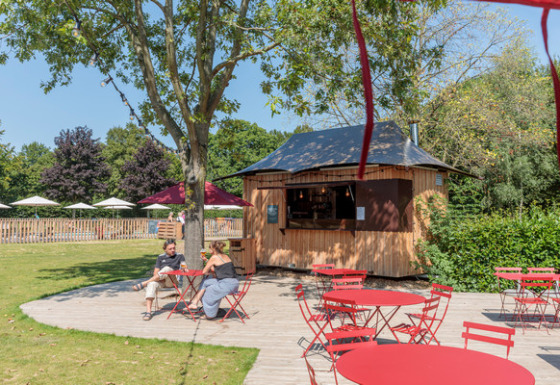 Zona caffè all’aperto con tavoli e sedie rosse accanto a una casetta in legno al Camping Valkenburg, Limburgo, Paesi Bassi.