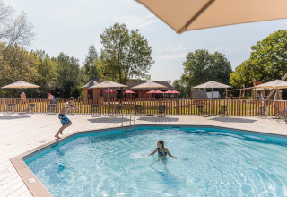 Children playing in the pool at Camping Valkenburg - Maastricht, surrounded by sun umbrellas and lush greenery.