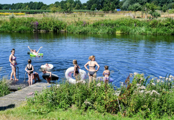 Children and adults enjoy swimming and playing with float rings by a lush riverside on a sunny day.