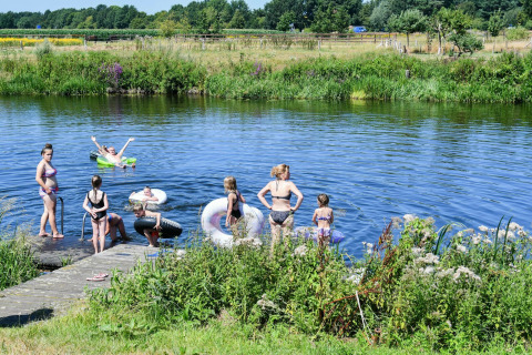Enfants et adultes nagent et jouent avec des bouées le long d’une rivière entourée de verdure au soleil.