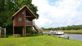 Boomhut op hout aan het water, groene grasvlakte en een passerende boot op de rivier.