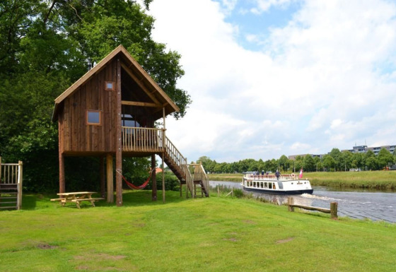 Cabane perchée en bois au bord d’une rivière, pelouse verte, banc de pique-nique et un bateau.