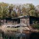 En moderne lodge ved en dam med springvand, omgivet af træer og natur på en solrig dag i skoven.