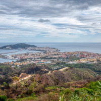 Vista de una ciudad costera junto al mar con colinas verdes, parque vacacional y opciones de glamping bajo cielo nublado.