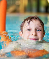Niño sonriente nada en la piscina con churro naranja en parque vacacional con glamping.