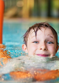 Smiling child swimming with orange noodle in a holiday park pool offering glamping accommodations.