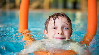 Niño sonriente nada en la piscina con churro naranja en parque vacacional con glamping.