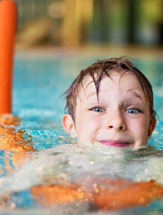 Niño sonriente nada en la piscina con churro naranja en parque vacacional con glamping.
