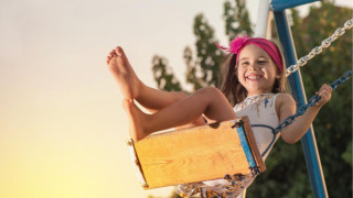 Niña feliz con diadema rosa juega en un columpio al atardecer en parque vacacional con glamping.