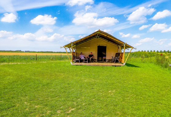 Two people relax on the porch of a safari tent with toilet, set in a green field under a blue sky.