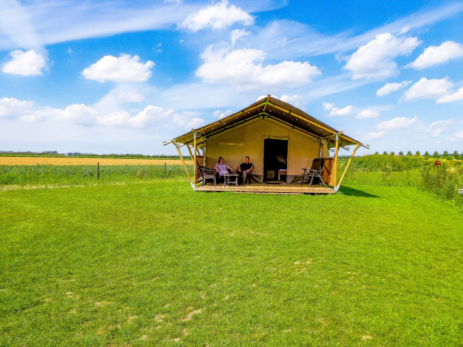 Zwei Personen entspannen vor einem Safarizelt mit Toilette auf einer grünen Wiese unter blauem Himmel.
