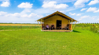 Two people relax on the porch of a safari tent with toilet, set in a green field under a blue sky.