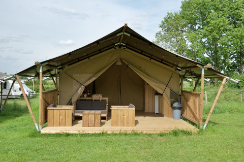 Safari tent with terrace, seating area, and toilet on grassy campsite, equipped with wooden furniture.
