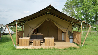 Safari tent with terrace, seating area, and toilet on grassy campsite, equipped with wooden furniture.