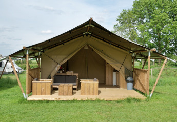 Tenda safari con veranda, area salotto e toilette su prato, dotata di mobili in legno stile campeggio.