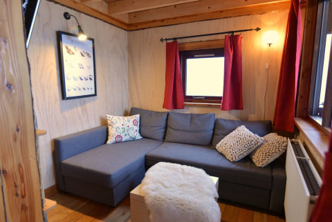 Knusse zithoek in de boomhut bij Camping de Koeksebelt, met grijze zetel en rode gordijnen.