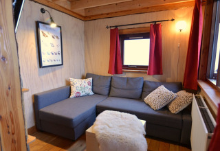 Gemütliches Wohnzimmer im Baumhaus auf Camping de Koeksebelt, mit grauem Sofa und roten Vorhängen.