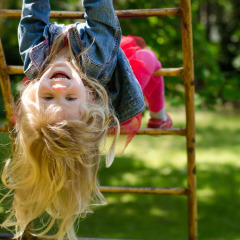 Happy girl hanging upside down on climbing bars outdoors in a sunny holiday park with glamping.