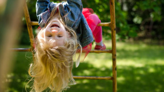 Niña feliz colgando boca abajo en barras de juego al aire libre en un parque vacacional con glamping.