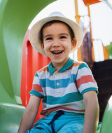 Niño sonriente con sombrero blanco sentado en un tobogán verde en un parque de vacaciones con glamping.