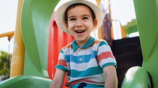 Niño sonriente con sombrero blanco sentado en un tobogán verde en un parque de vacaciones con glamping.